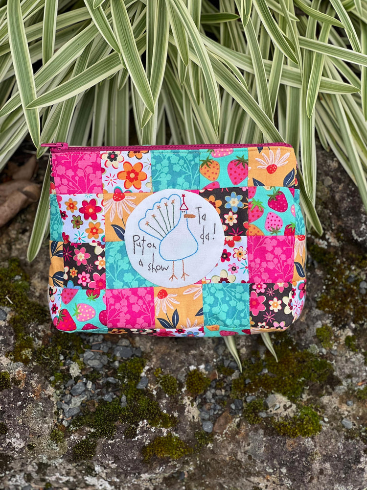 Colorful patchwork pouch with a brand logo on a textured surface with plants in the background